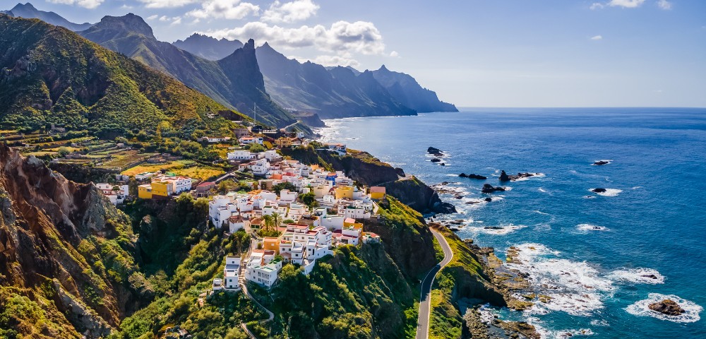A coastal village with white and colorful houses atop lush cliffs overlooks a rocky shoreline and blue ocean. Mountainous terrain rises in the background under a clear sky.