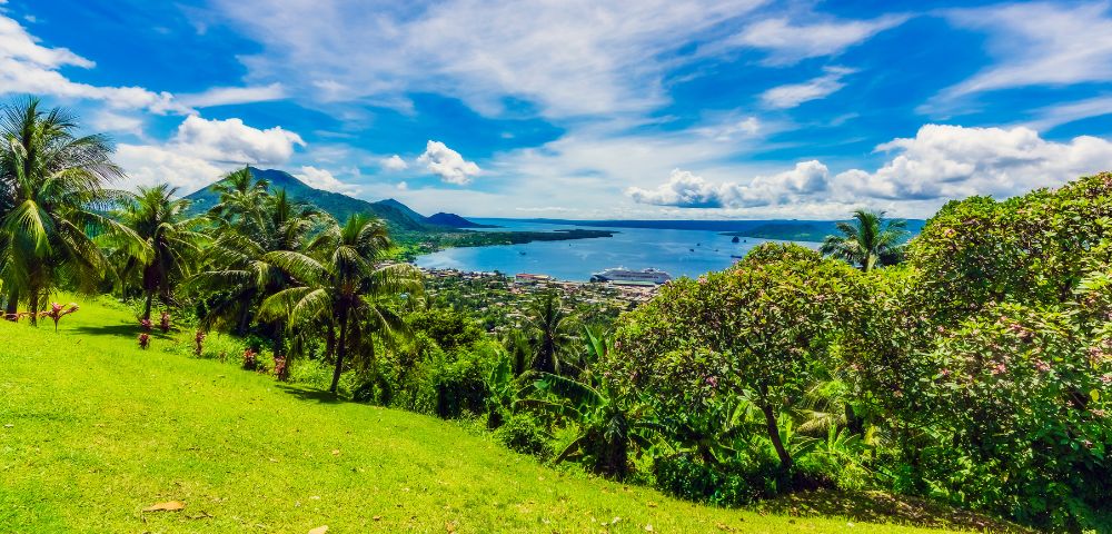 Tropical landscape with lush green hills, palm trees, and a vivid blue sea under a partly cloudy sky. Relaxing and vibrant coastal scene.