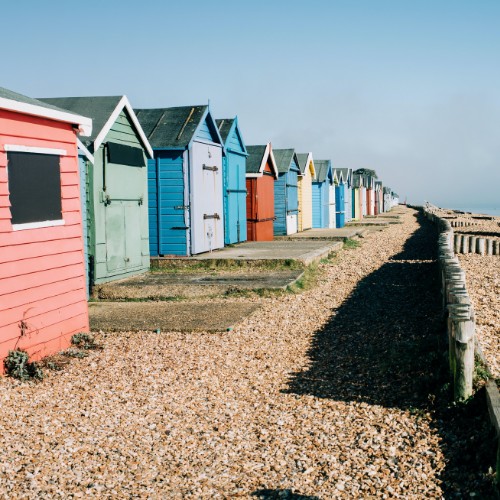 Colorful beach huts line a pebbled shore, some with red, blue, or green hues. The calm sea and clear sky create a tranquil atmosphere.
