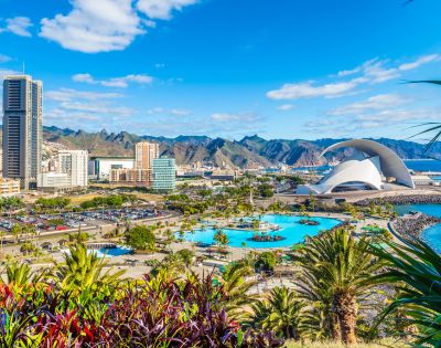 A vibrant coastal cityscape with tall modern buildings, a unique curved architectural structure by the sea, lush greenery, and distant mountains under a blue sky.
