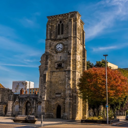 Historic stone church tower with a clock under a clear blue sky, surrounded by trees with autumn foliage. The scene evokes a calm, timeless feel.