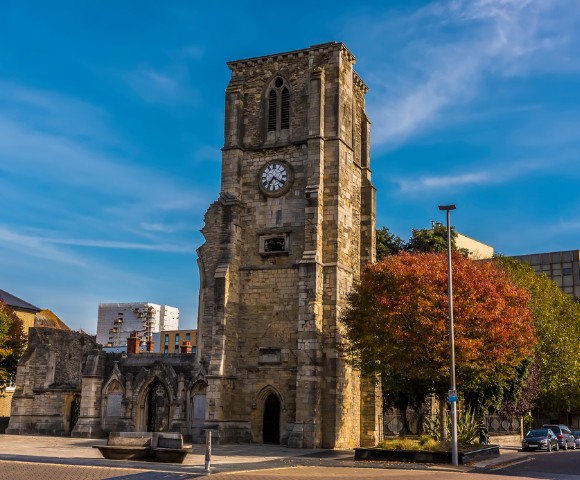 Historic stone church tower with a clock under a clear blue sky, surrounded by trees with autumn foliage. The scene evokes a calm, timeless feel.