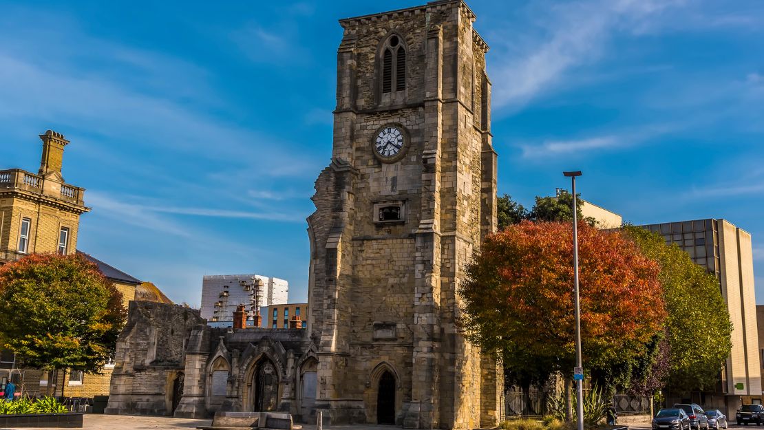 Historic stone church tower with a clock under a clear blue sky, surrounded by trees with autumn foliage. The scene evokes a calm, timeless feel.