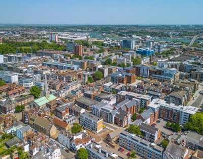 Aerial view of a cityscape on a clear day, featuring densely packed buildings of various heights and architectural styles, surrounded by green spaces.