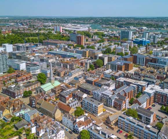 Aerial view of a cityscape on a clear day, featuring densely packed buildings of various heights and architectural styles, surrounded by green spaces.