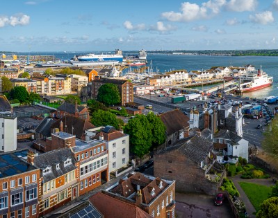 A vibrant port cityscape featuring colorful historic buildings in the foreground, a bustling harbor with a large ferry and clear blue sky.