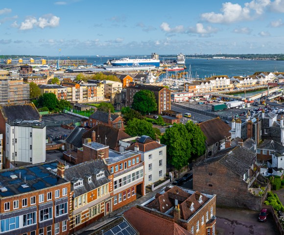 A vibrant port cityscape featuring colorful historic buildings in the foreground, a bustling harbor with a large ferry and clear blue sky.