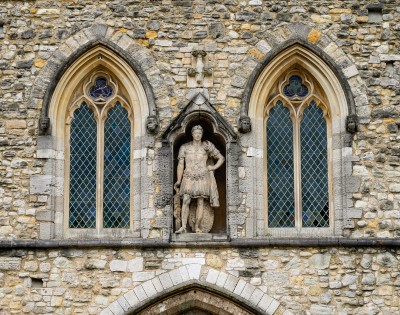 Facade of an old stone building with two arched windows showcasing intricate lattice designs. A central statue depicts a historical figure in armor, adding a majestic feel.