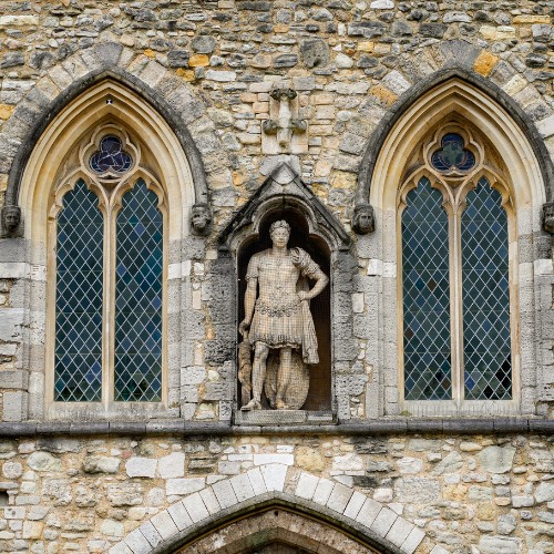 Facade of an old stone building with two arched windows showcasing intricate lattice designs. A central statue depicts a historical figure in armor, adding a majestic feel.
