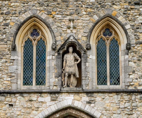 Facade of an old stone building with two arched windows showcasing intricate lattice designs. A central statue depicts a historical figure in armor, adding a majestic feel.