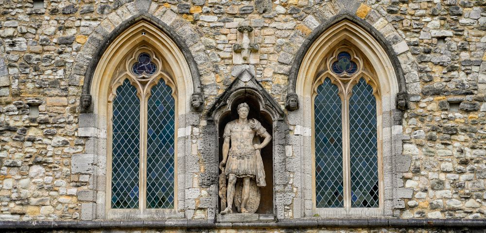 Facade of an old stone building with two arched windows showcasing intricate lattice designs. A central statue depicts a historical figure in armor, adding a majestic feel.