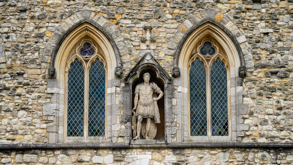 Facade of an old stone building with two arched windows showcasing intricate lattice designs. A central statue depicts a historical figure in armor, adding a majestic feel.