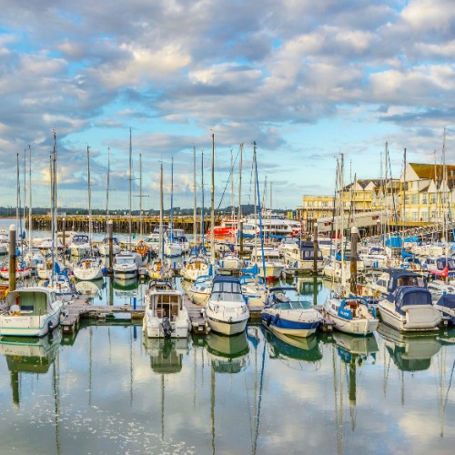 A picturesque harbor filled with various docked sailboats reflecting on calm water. The scene is under a partly cloudy sky, with charming buildings in the background.
