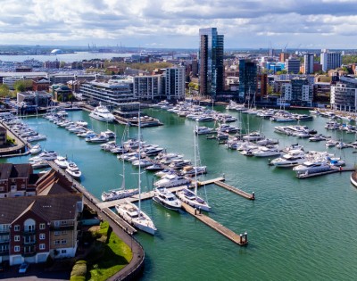 Aerial view of a marina filled with white yachts and boats, surrounded by modern buildings under a partly cloudy sky. The scene conveys a sense of tranquility.