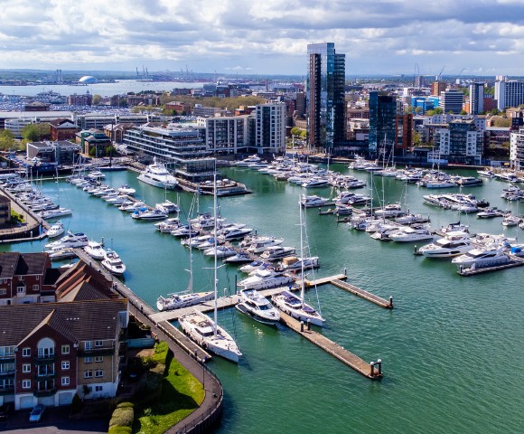 Aerial view of a marina filled with white yachts and boats, surrounded by modern buildings under a partly cloudy sky. The scene conveys a sense of tranquility.