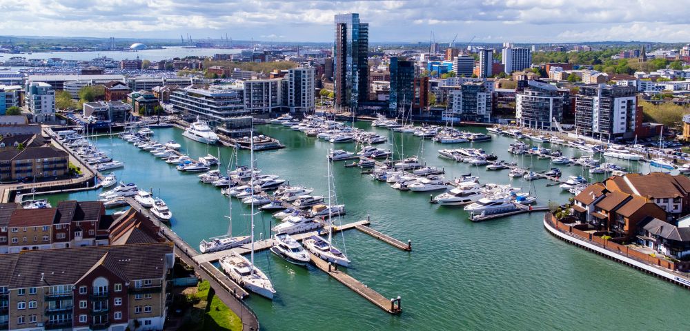 Aerial view of a marina filled with white yachts and boats, surrounded by modern buildings under a partly cloudy sky. The scene conveys a sense of tranquility.