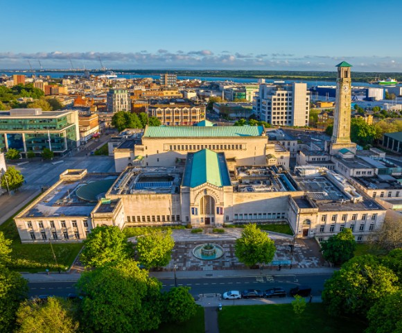 Aerial view of a cityscape featuring a large building with a green roof and clock tower. Surrounding it are trees, roads, and various urban structures. Clear blue sky enhances the vibrant setting.