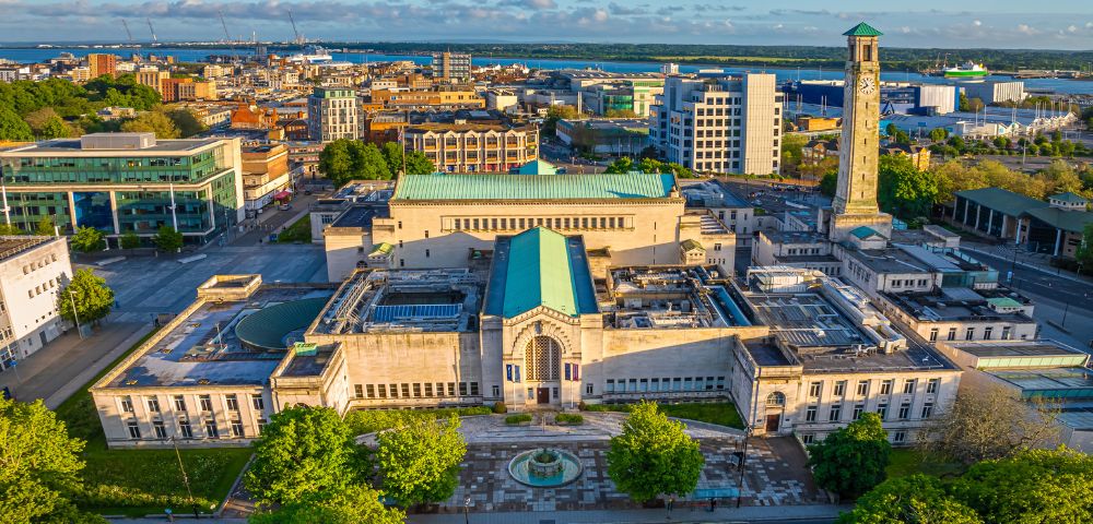 Aerial view of a cityscape featuring a large building with a green roof and clock tower. Surrounding it are trees, roads, and various urban structures. Clear blue sky enhances the vibrant setting.