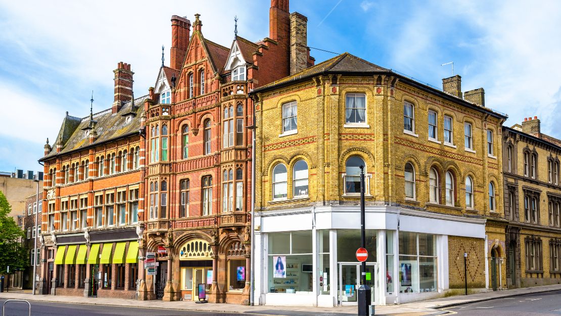 Historic street corner featuring colorful Victorian buildings with ornate brickwork and large arched windows, set against a blue sky.