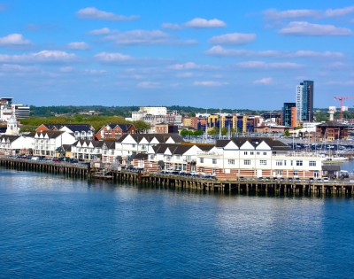Coastal cityscape with a row of white waterfront buildings under a blue sky with scattered clouds. A red and white ferry is docked nearby.