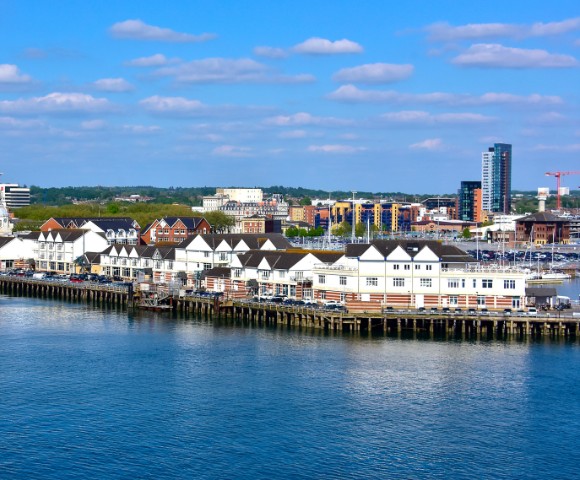 Coastal cityscape with a row of white waterfront buildings under a blue sky with scattered clouds. A red and white ferry is docked nearby.