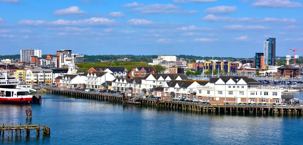 Coastal cityscape with a row of white waterfront buildings under a blue sky with scattered clouds. A red and white ferry is docked nearby.