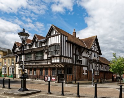 A historic Tudor-style building with exposed timber framing and steep gabled roofs stands under a partly cloudy sky, exuding a nostalgic charm.