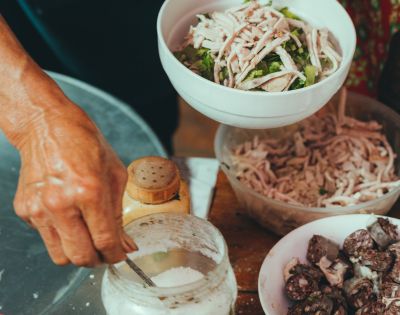 Close-up of a person preparing food, mixing shredded chicken with herbs in a white bowl. Ingredients like a spice jar, flour, and sausages are nearby, creating a rustic, culinary scene.