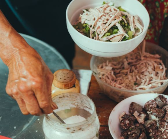 Close-up of a person preparing food, mixing shredded chicken with herbs in a white bowl. Ingredients like a spice jar, flour, and sausages are nearby, creating a rustic, culinary scene.
