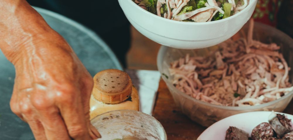 Close-up of a person preparing food, mixing shredded chicken with herbs in a white bowl. Ingredients like a spice jar, flour, and sausages are nearby, creating a rustic, culinary scene.
