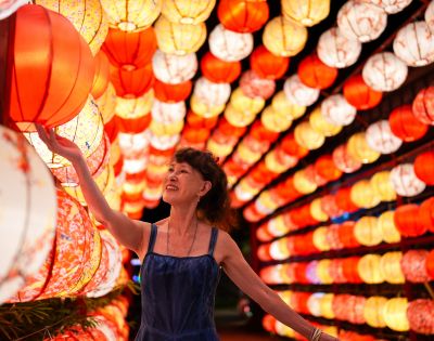 A woman in a blue dress smiles while touching a red lantern. She's surrounded by a vibrant display of red and yellow lanterns, creating a festive atmosphere.