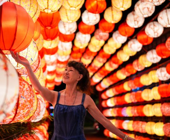 A woman in a blue dress smiles while touching a red lantern. She's surrounded by a vibrant display of red and yellow lanterns, creating a festive atmosphere.