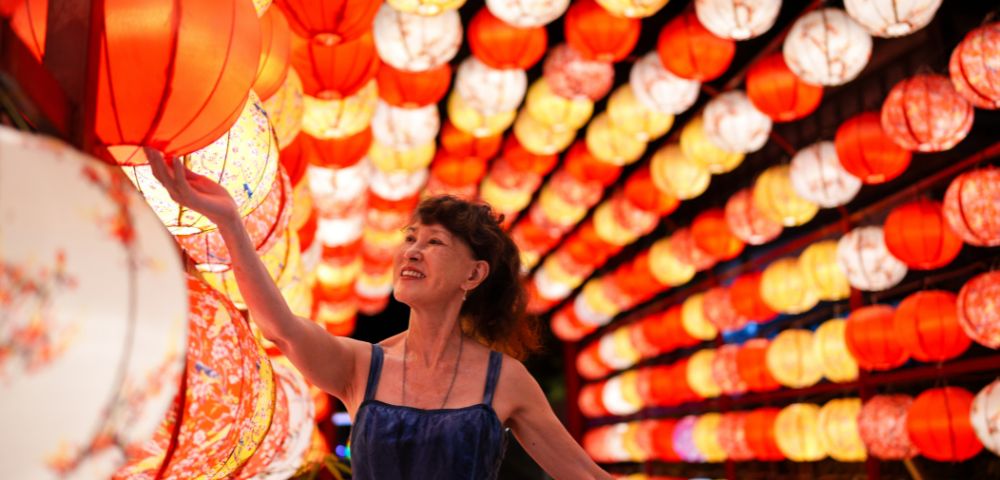 A woman in a blue dress smiles while touching a red lantern. She's surrounded by a vibrant display of red and yellow lanterns, creating a festive atmosphere.