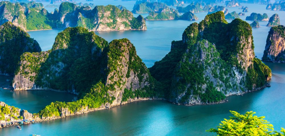 Scenic view of Ha Long Bay, Vietnam, with towering limestone islands covered in green foliage, surrounded by tranquil blue water under a clear sky.