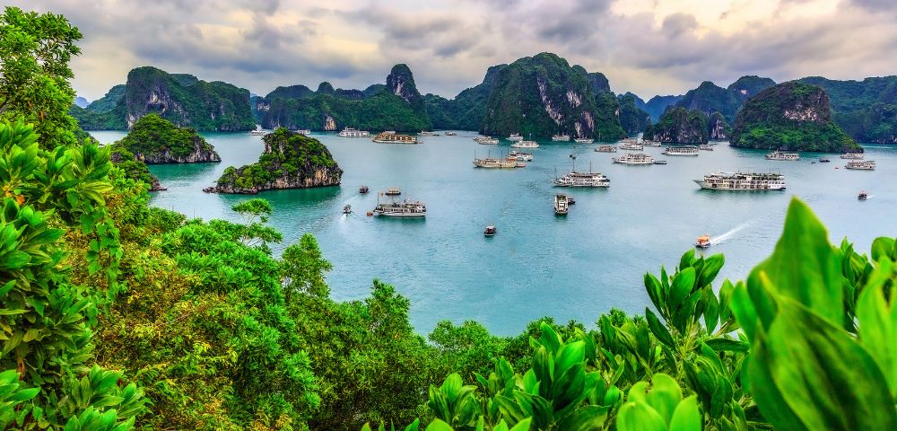Scenic view of Ha Long Bay with emerald waters, lush green islands, and boats gently sailing beneath a cloudy sky, conveying tranquility and natural beauty.