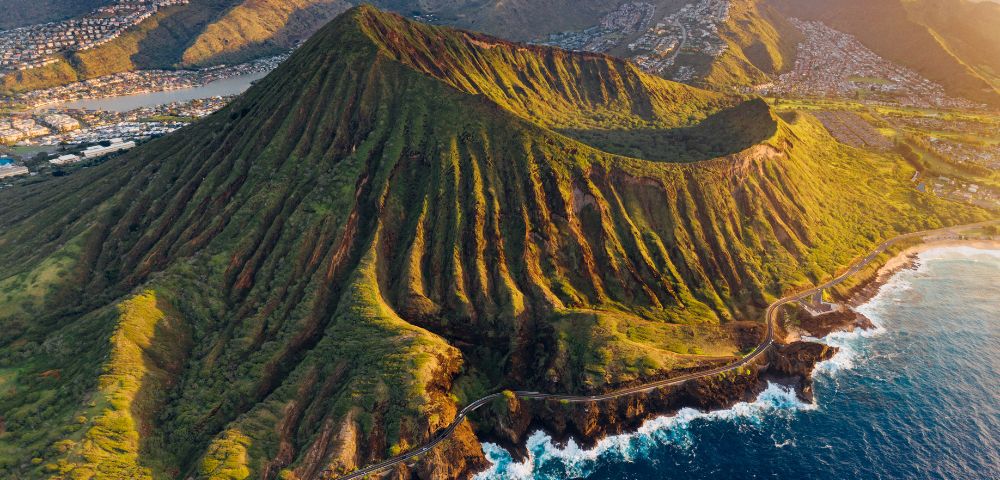 Aerial view of a lush, green volcanic mountain with deep ridges on a coastline. Blue ocean waves crash against the rocky shore. Nearby, a town sits in the valley.