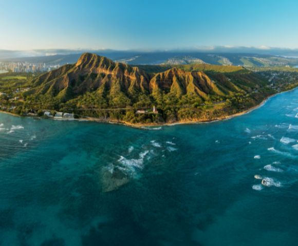 Aerial view of Diamond Head, a dormant volcanic crater on Oahu, Hawaii, surrounded by turquoise ocean, with Honolulu's skyline in the background.