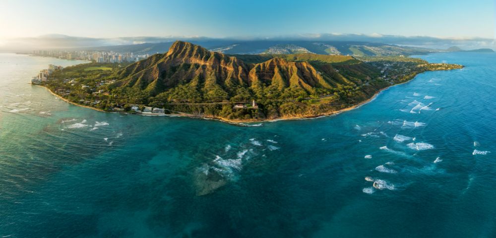 Aerial view of Diamond Head, a dormant volcanic crater on Oahu, Hawaii, surrounded by turquoise ocean, with Honolulu's skyline in the background.
