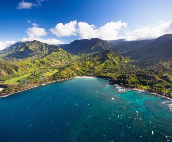 Aerial view of a lush, green mountain range beside a pristine, turquoise bay. The sky is clear with a few clouds, evoking a serene and beautiful scene.