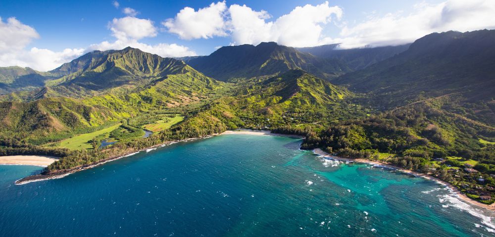 Aerial view of a lush, green mountain range beside a pristine, turquoise bay. The sky is clear with a few clouds, evoking a serene and beautiful scene.