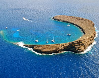 Aerial view of a crescent-shaped island surrounded by vibrant blue ocean. Boats are anchored in the calm, clear waters within the island's embrace.