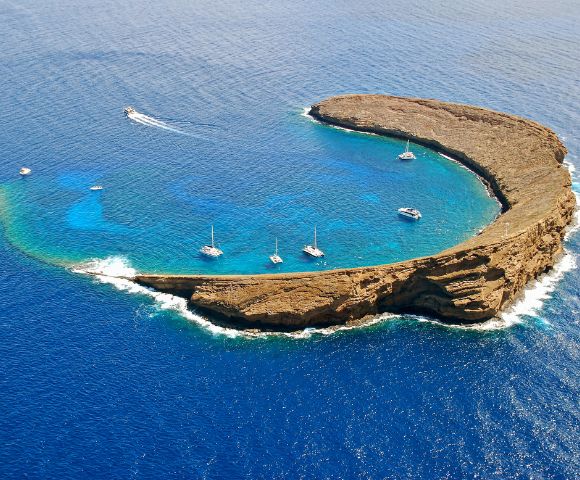 Aerial view of a crescent-shaped island surrounded by vibrant blue ocean. Boats are anchored in the calm, clear waters within the island's embrace.