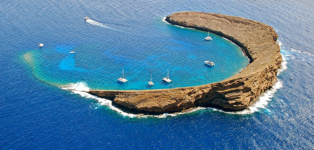 Aerial view of a crescent-shaped island surrounded by vibrant blue ocean. Boats are anchored in the calm, clear waters within the island's embrace.