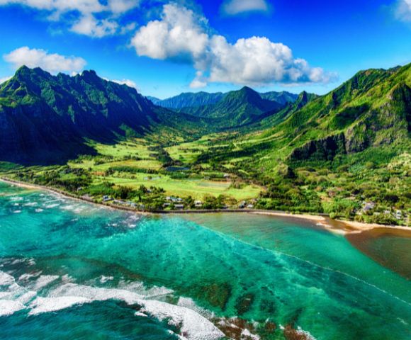 Aerial view of a lush, green mountainous landscape with clear turquoise ocean waves lapping against a sandy coastline under a partly cloudy blue sky.