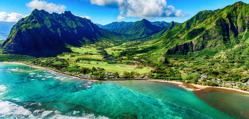 Aerial view of a lush, green mountainous landscape with clear turquoise ocean waves lapping against a sandy coastline under a partly cloudy blue sky.