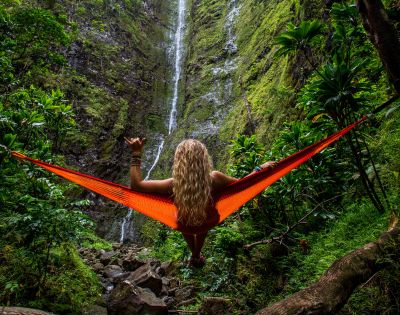 A person with long blonde hair relaxes in an orange hammock facing a tall waterfall surrounded by lush green foliage, conveying tranquility and adventure.