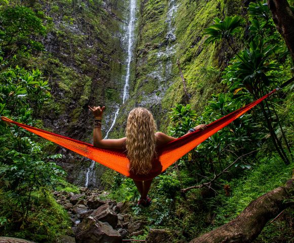 A person with long blonde hair relaxes in an orange hammock facing a tall waterfall surrounded by lush green foliage, conveying tranquility and adventure.