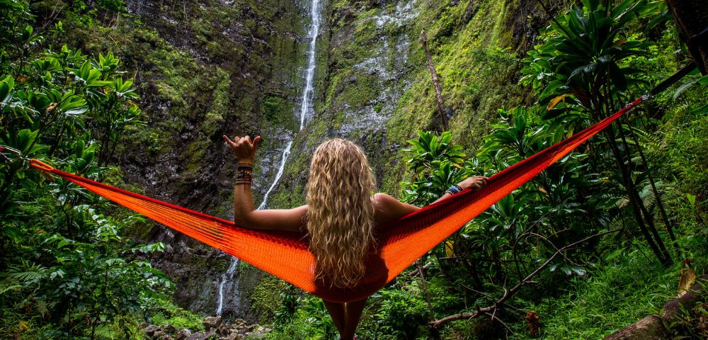 A person with long blonde hair relaxes in an orange hammock facing a tall waterfall surrounded by lush green foliage, conveying tranquility and adventure.