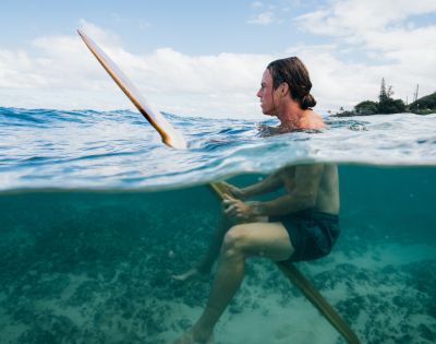 A person with long hair and wearing dark shorts floats on a surfboard in clear blue ocean water. The horizon shows a partly cloudy sky and distant trees, conveying a serene and adventurous mood.