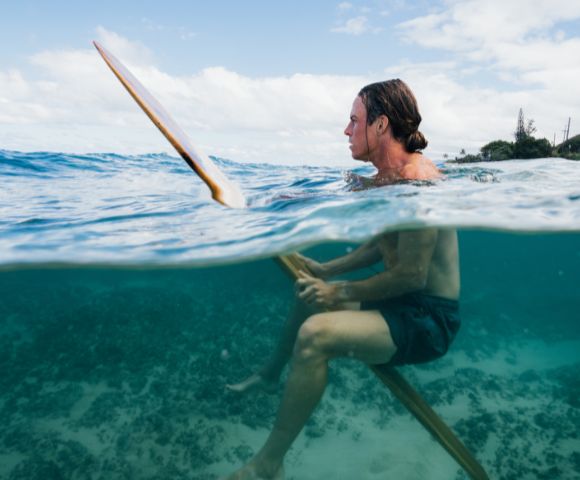 A person with long hair and wearing dark shorts floats on a surfboard in clear blue ocean water. The horizon shows a partly cloudy sky and distant trees, conveying a serene and adventurous mood.